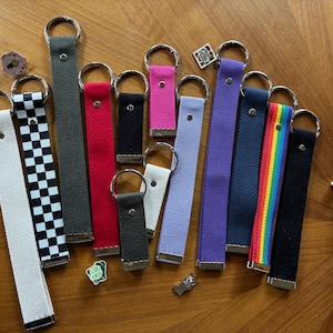 May include: A collection of fabric wristlet keychains in various colors and patterns, including solid white, black, red, gray, purple, pink, light blue, a black and white checkerboard print, and a rainbow striped design, each with a metal ring.
