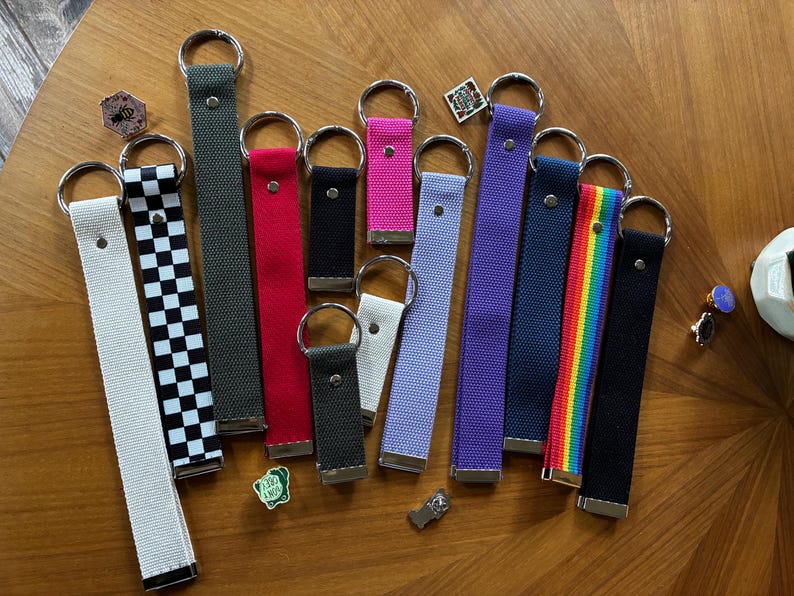 May include: A collection of fabric wristlet keychains in various colors and patterns, including solid white, black, red, gray, purple, pink, light blue, a black and white checkerboard print, and a rainbow striped design, each with a metal ring.