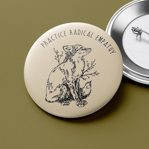 May include: A round, tan button with a black illustration of a fox and sprigs. The text "PRACTICE RADICAL EMPATHY" is printed in a semi-circle around the fox. A second button is partially visible.