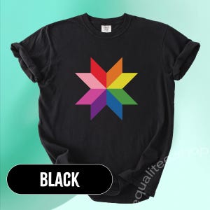 May include: Black t-shirt featuring a colorful starburst design in rainbow colors. The shirt has rolled-up sleeves and the word "BLACK" in a black rectangle at the bottom. The shirt is a comfortable, casual style.