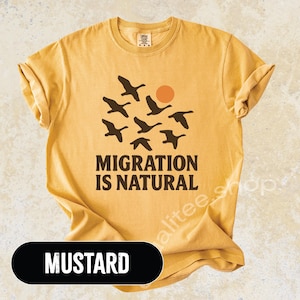 May include: Mustard-colored t-shirt with a graphic of flying birds and a sun. The text "MIGRATION IS NATURAL" is printed below the birds in a bold font. The shirt is made of a soft material.