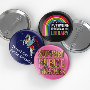 May include: A collection of round pinback buttons with various designs. One button features a rainbow and the text "EVERYONE BELONGS AT THE LIBRARY." Another says "Defend Our Public Libraries" with a knight on horseback. A third says "Support Public Libraries."