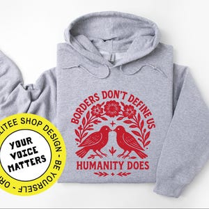 Pode incluir: Um moletom cinza claro com um gr&aacute;fico vermelho que diz "Borders Don't Define Us, Humanity Does". O design inclui dois p&aacute;ssaros e elementos florais. Um c&iacute;rculo amarelo com o texto "Your Voice Matters" est&aacute; no canto inferior esquerdo.