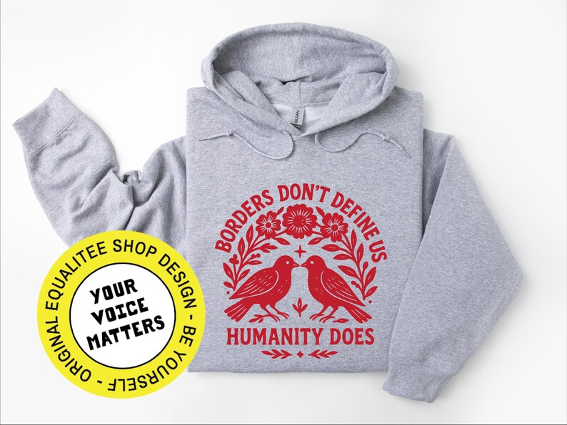 Pode incluir: Um moletom cinza claro com um gr&aacute;fico vermelho que diz "Borders Don't Define Us, Humanity Does". O design inclui dois p&aacute;ssaros e elementos florais. Um c&iacute;rculo amarelo com o texto "Your Voice Matters" est&aacute; no canto inferior esquerdo.