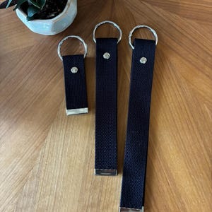 May include: Three black fabric key fobs of varying lengths, each featuring a silver-toned metal split ring at the top, a silver-toned metal rivet near the top, and a silver-toned metal end cap at the bottom, arranged in a vertical row.