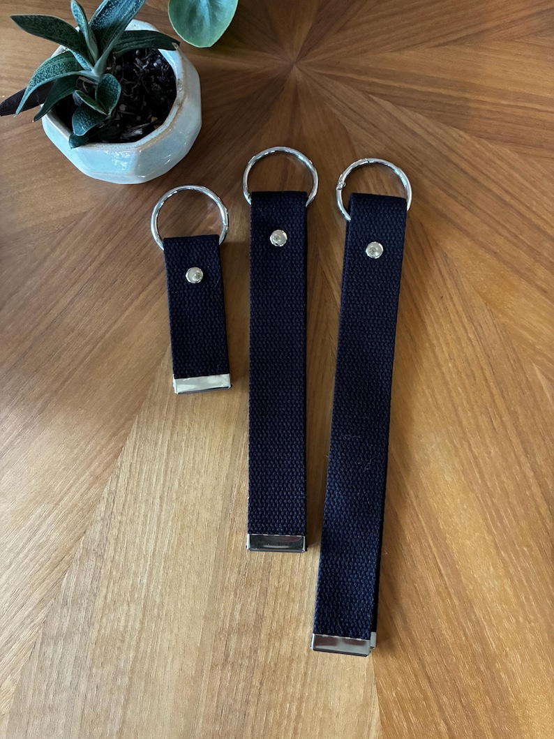 May include: Three black fabric key fobs of varying lengths, each featuring a silver-toned metal split ring at the top, a silver-toned metal rivet near the top, and a silver-toned metal end cap at the bottom, arranged in a vertical row.