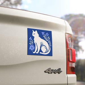 May include: A white cat silhouette on a blue square, surrounded by floral elements. The design is affixed to the back of a white vehicle. The cat is sitting, with a detailed floral pattern on its body. The background is a light blue.
