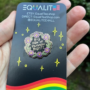 Pu&ograve; includere: Una carta nera con un arcobaleno in basso e la scritta "EQUALITE" in alto. Una spilla con la scritta "Big Fan of Human Rights" &egrave; al centro. La carta riporta anche il nome del negozio Etsy e l'handle dei social media.