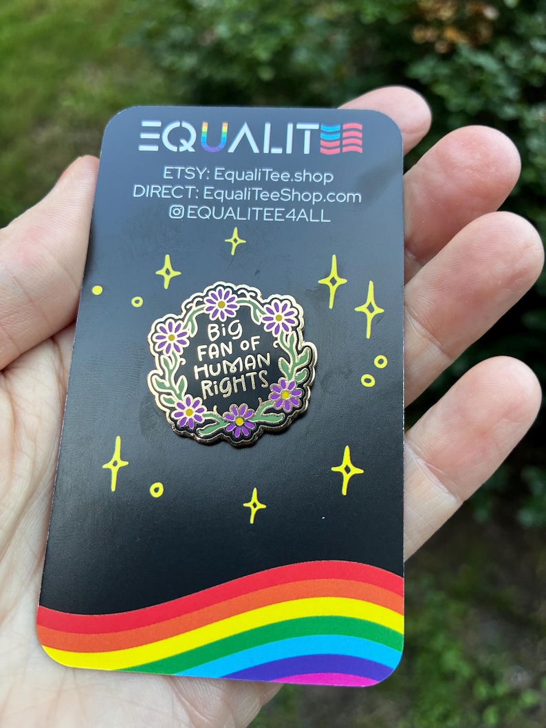 Pu&ograve; includere: Una carta nera con un arcobaleno in basso e la scritta "EQUALITE" in alto. Una spilla con la scritta "Big Fan of Human Rights" &egrave; al centro. La carta riporta anche il nome del negozio Etsy e l'handle dei social media.