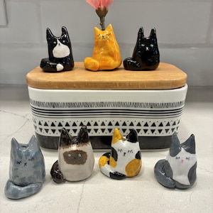 May include: Seven small ceramic cat figurines in various colors and patterns, including black, white, orange, gray, and calico, arranged around a rectangular white and black patterned container with a wooden lid holding a pink flower.
