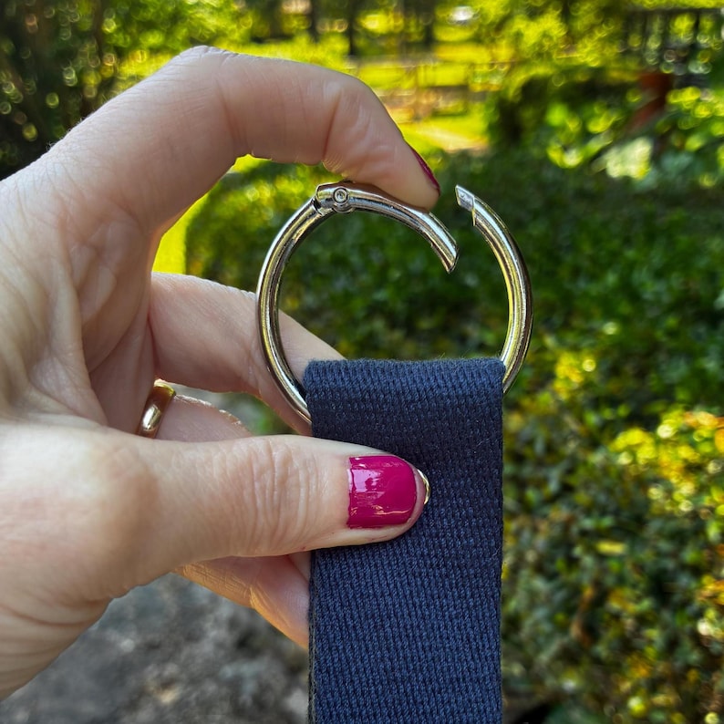 May include: A circular silver-toned metal spring gate ring attached to a dark navy blue woven fabric strap. The metal ring features a hinged opening mechanism that allows the loop to be opened and closed, securely holding the flat textile webbing.