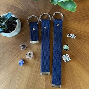 May include: Three navy blue fabric key fobs of varying lengths, each featuring a silver metal ring at the top and a silver metal clamp at the bottom. Several small decorative enamel pins are scattered around the key fobs, including designs of a bee, a frog, and a national park emblem.