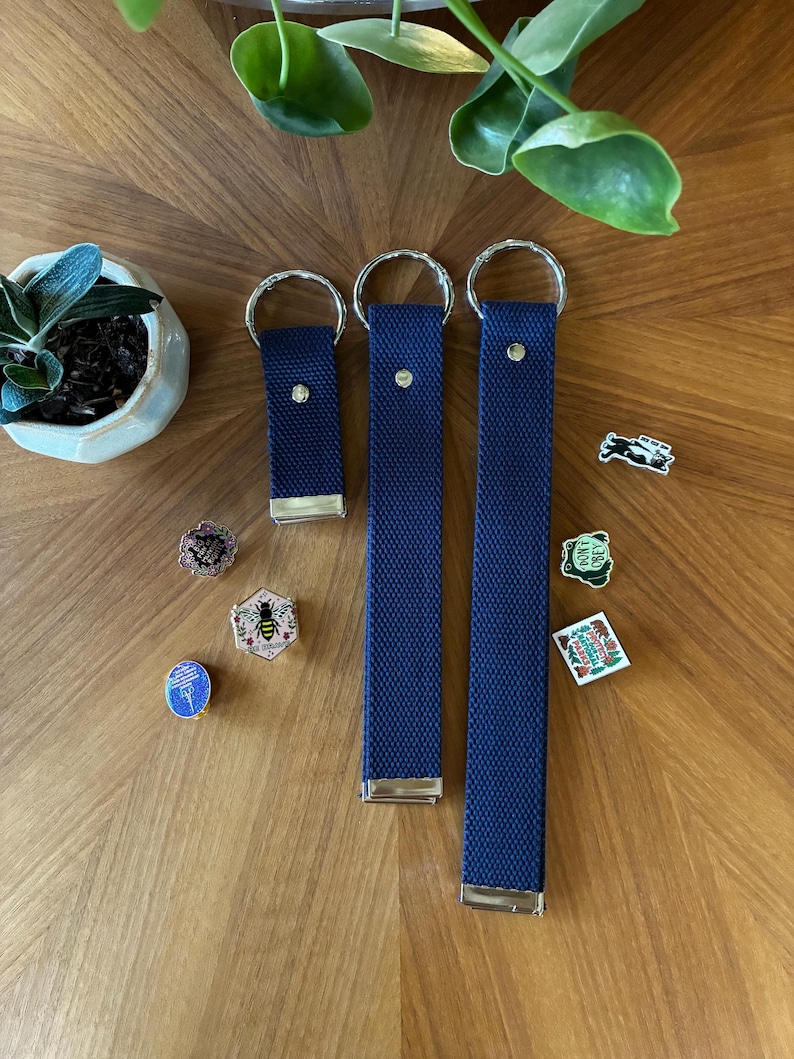 May include: Three navy blue fabric key fobs of varying lengths, each featuring a silver metal ring at the top and a silver metal clamp at the bottom. Several small decorative enamel pins are scattered around the key fobs, including designs of a bee, a frog, and a national park emblem.