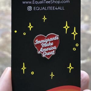 Pu&ograve; includere: Una carta nera con il testo "EQUALITE" e le informazioni sul sito web. Una spilla a forma di cuore rosso con le parole "Immigrants Make America Great" &egrave; al centro. Esplosioni di stelle gialle e un bordo arcobaleno completano il design.