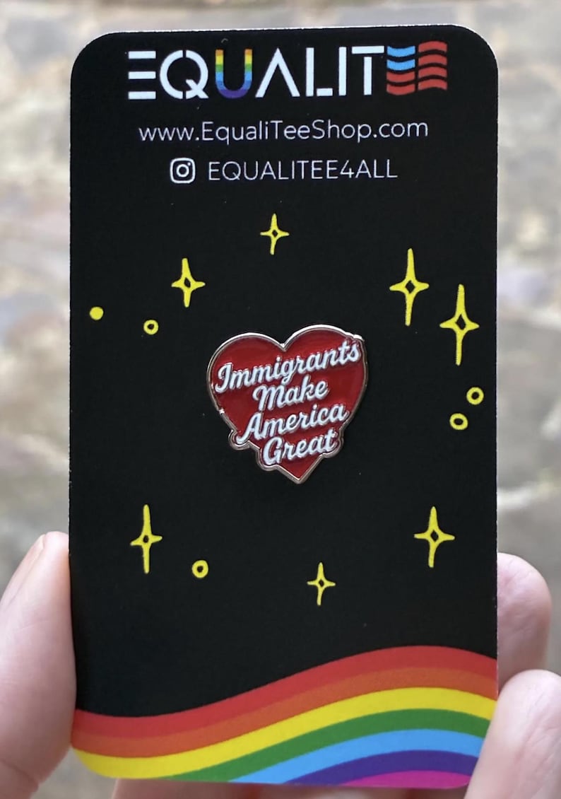 Pu&ograve; includere: Una carta nera con il testo "EQUALITE" e le informazioni sul sito web. Una spilla a forma di cuore rosso con le parole "Immigrants Make America Great" &egrave; al centro. Esplosioni di stelle gialle e un bordo arcobaleno completano il design.