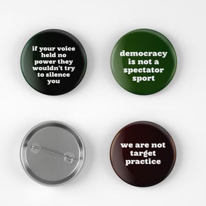 May include: Four round buttons with white text on colored backgrounds. The buttons are black, dark green, and brown. The text on the buttons reads: "if your voice held no power they wouldn't try to silence you", "democracy is not a spectator sport", and "we are not target practice".
