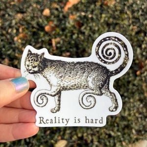 May include: A white sticker featuring a stylized cat illustration in black and gray. The cat has a swirled tail and paws. The text "Reality is hard" is printed below the cat in a serif font. The sticker is held by a hand.