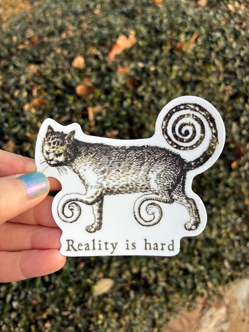 May include: A white sticker featuring a stylized cat illustration in black and gray. The cat has a swirled tail and paws. The text "Reality is hard" is printed below the cat in a serif font. The sticker is held by a hand.