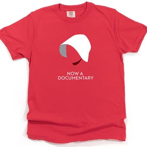 Puede incluir: Camiseta roja con una silueta blanca de una mujer con una capucha blanca y el texto "NOW A DOCUMENTARY".