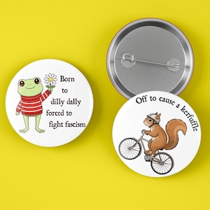 May include: Three round white pinback buttons with cartoon illustrations and text. One button features a frog in a red and white striped sweater holding a daisy, with the text "Born to dilly dally forced to fight fascism". Another shows a squirrel riding a bicycle with the text "Off to cause a kerfuffle".