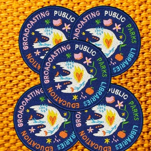 Pode incluir: Cinco patches bordados em azul marinho com as palavras "Broadcasting Public Education Parks Libraries" na borda. Cada patch apresenta um p&aacute;ssaro branco com um cora&ccedil;&atilde;o amarelo e laranja, rodeado por flores e estrelas. Os patches est&atilde;o em um fundo laranja texturizado.