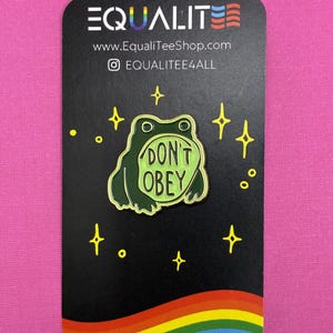 May include: A green frog pin with the words "DON'T OBEY" on a black card with a rainbow and stars. The card also has the text "EQUALITE" and a website address.