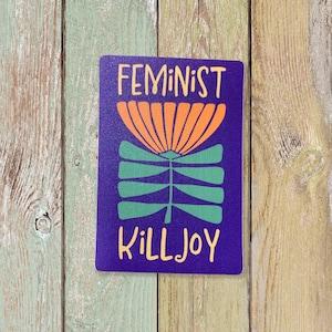 May include: A purple sticker with a stylized orange flower and green stem. The text "Feminist Killjoy" is written in white.