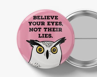 Pode incluir: Um botão redondo rosa com o texto "BELIEVE YOUR EYES, NOT THEIR LIES." apresenta uma ilustração de desenho animado de uma coruja. A coruja tem olhos amarelos e penas pretas e brancas. O botão tem uma parte traseira de metal.