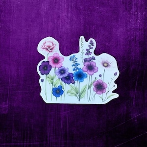 May include: A sticker featuring a cluster of purple, pink, and blue flowers with green stems and leaves. The flowers are arranged in a loose, organic pattern, creating a whimsical and delicate design.