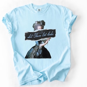 May include: Light blue t-shirt featuring a vintage portrait of a woman holding a sign that reads "Let Them Eat Cake." The graphic is a distressed design, adding a unique touch to the shirt. The shirt has short sleeves and a crew neck.