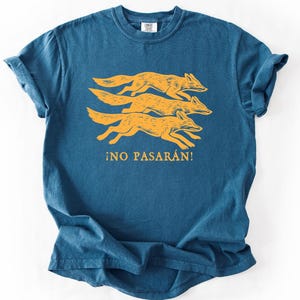 May include: A teal-colored t-shirt featuring a design of three running foxes in a golden-yellow hue. Below the foxes, the Spanish phrase "&iexcl;NO PASAR&Aacute;N!" is printed in the same golden-yellow color. The shirt has a classic crew neck and short sleeves.
