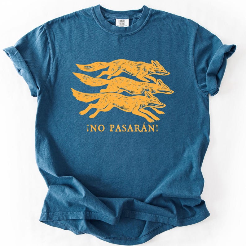 May include: A teal-colored t-shirt featuring a design of three running foxes in a golden-yellow hue. Below the foxes, the Spanish phrase "&iexcl;NO PASAR&Aacute;N!" is printed in the same golden-yellow color. The shirt has a classic crew neck and short sleeves.
