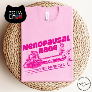 May include: Pink t-shirt with a pink graphic of a woman riding a chainsaw with the text "Menopausal Rage The Musical".