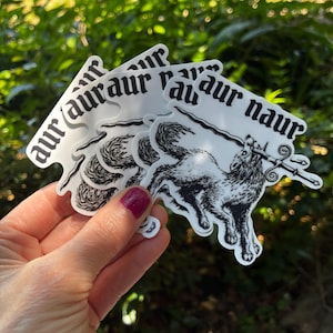 May include: A stack of white stickers with a black illustration of a wolf holding a sword. The stickers have the text "aur naur" in a gothic font. The stickers are held in front of a green leafy background.