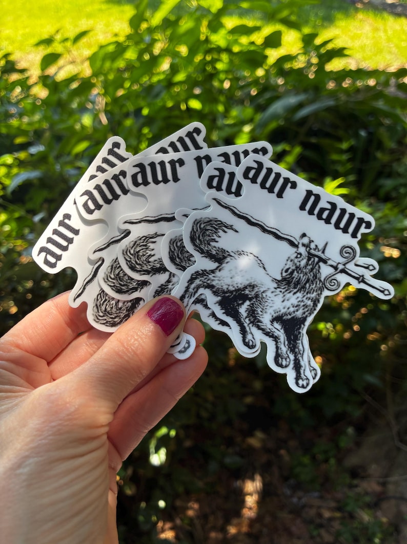 May include: A stack of white stickers with a black illustration of a wolf holding a sword. The stickers have the text "aur naur" in a gothic font. The stickers are held in front of a green leafy background.