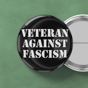 May include: A black button with the white text "VETERAN AGAINST FASCISM" is displayed. The button is glossy and round, with a silver metal edge. Another button is partially visible in the background.
