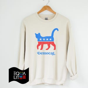 K&ouml;nnte beinhalten: Ein cremefarbener Pullover mit einer blauen und roten Katzensilhouette, die ein Design tr&auml;gt, das an die amerikanische Flagge erinnert. Der Text "Democat" ist unter der Katze aufgedruckt.