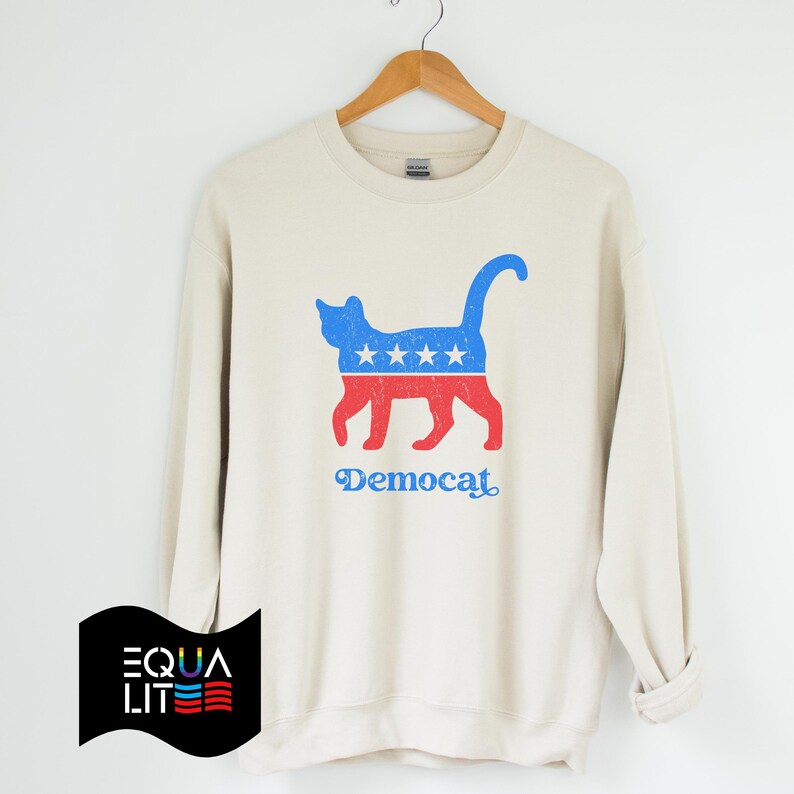 K&ouml;nnte beinhalten: Ein cremefarbener Pullover mit einer blauen und roten Katzensilhouette, die ein Design tr&auml;gt, das an die amerikanische Flagge erinnert. Der Text "Democat" ist unter der Katze aufgedruckt.