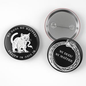 May include: Set of three round buttons. Two black buttons feature white illustrations and text. One button shows a cat with a snake and the text "NO GODS NO MASTERS NI DIEU NI MAÎTRE". The other button shows a snake eating its tail. The third button is silver.