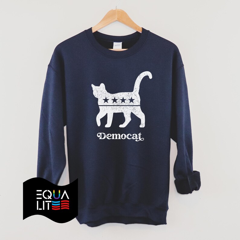 K&ouml;nnte beinhalten: Ein marineblauer Pullover mit einer wei&szlig;en Silhouette einer Katze mit Sternen auf dem R&uuml;cken. Der Text "Democat" ist unter der Katze aufgedruckt.