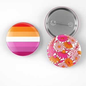 May include: Two round pinback buttons. One features the lesbian pride flag with horizontal stripes of red, orange, white, pink, and magenta. The other button has a floral design with pink, orange, and white flowers.