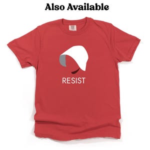 Puede incluir: Camiseta roja con una silueta blanca de una mujer con una capucha blanca y la palabra "RESIST" impresa en blanco debajo de la silueta.