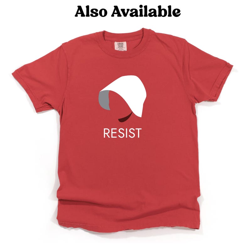 Puede incluir: Camiseta roja con una silueta blanca de una mujer con una capucha blanca y la palabra "RESIST" impresa en blanco debajo de la silueta.