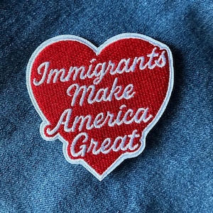 May include: A red heart-shaped patch with white embroidered text that reads "Immigrants Make America Great." The patch has a white border and is set against a blue denim background. The patch is likely designed to be sewn or ironed onto clothing or accessories.