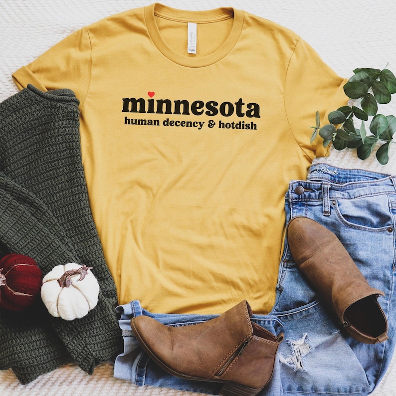 May include: A yellow t-shirt with the text "minnesota human decency & hotdish" printed on it. The shirt is laid out on a white surface with a pair of brown boots, blue jeans, and a green sweater.