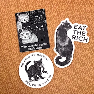 May include: A collection of black and white cat stickers with various designs. One sticker features multiple cats with the text "We're all in this together. Like hostages." Another says "Eat the Rich." A third sticker shows a cat with a snake and the text "No Gods No Masters."