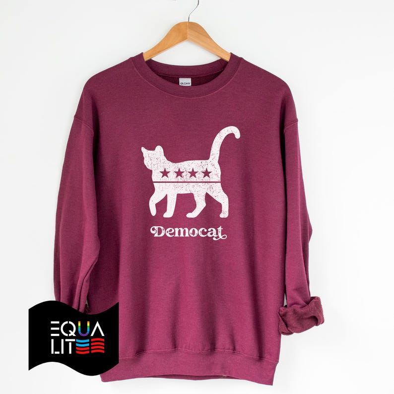 K&ouml;nnte beinhalten: Ein weinroter Pullover mit einer wei&szlig;en Silhouette einer Katze mit Sternen auf dem R&uuml;cken. Der Text "Democat" ist unter der Katze aufgedruckt.