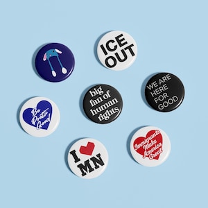 May include: A collection of seven round buttons with various designs and text on a light blue background. The buttons feature phrases like "ICE OUT," "We Are Here For Good," and "Immigrants Make America Great." Other buttons have heart and bunny designs.
