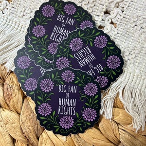 May include: A stack of round stickers with a black background and a floral wreath design. The stickers have the text "BIG FAN OF HUMAN RIGHTS" in white. The floral wreath features purple flowers and green leaves.