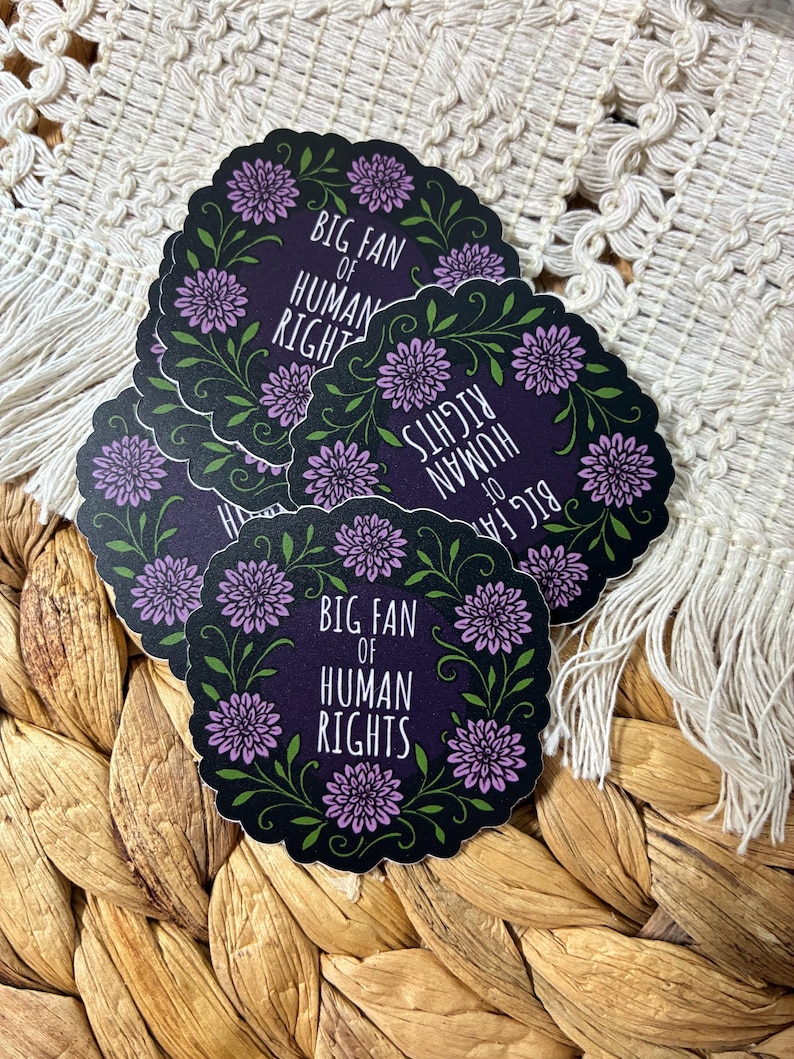 May include: A stack of round stickers with a black background and a floral wreath design. The stickers have the text "BIG FAN OF HUMAN RIGHTS" in white. The floral wreath features purple flowers and green leaves.